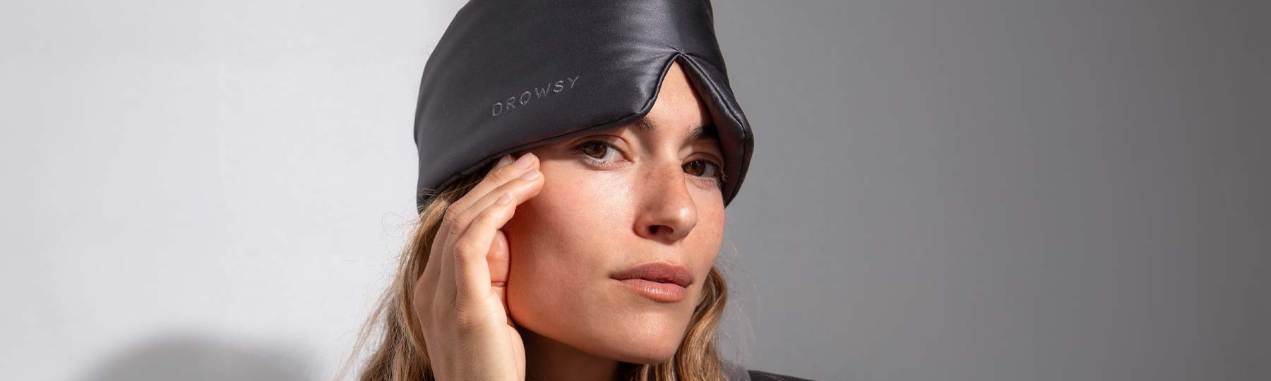A woman with long, light brown hair wears a black sleep mask labeled DROWSY on her head, touching the side of her face with one hand against a neutral background.