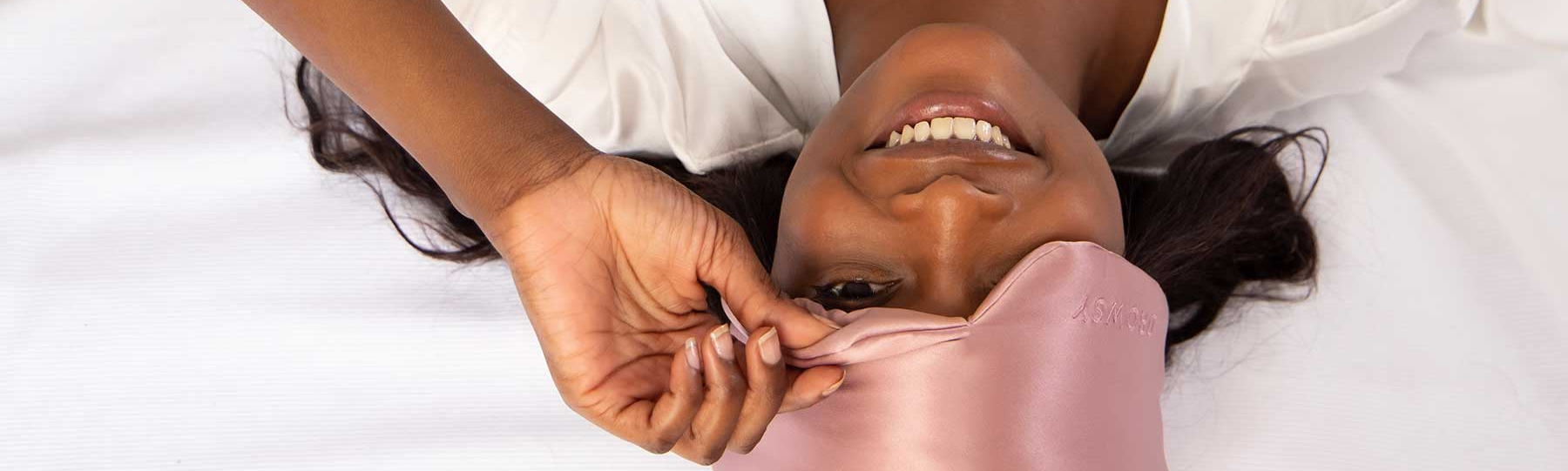 A woman lies on her back smiling, wearing a white top and adjusting a pink satin sleep mask over her eyes while resting on a white bed.