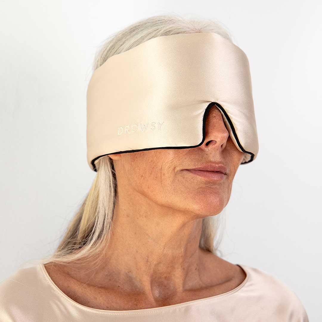An older woman with long gray hair wears a large, light gold satin sleep mask that covers her eyes and ears. The mask has DROWSY written on it and she is dressed in a matching top.