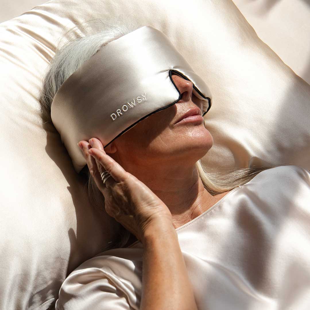 A woman with long gray hair lies on a satin pillow, wearing a shiny silk eye mask that says DROWSY. She is dressed in a matching satin top and gently touches the mask with her hand.