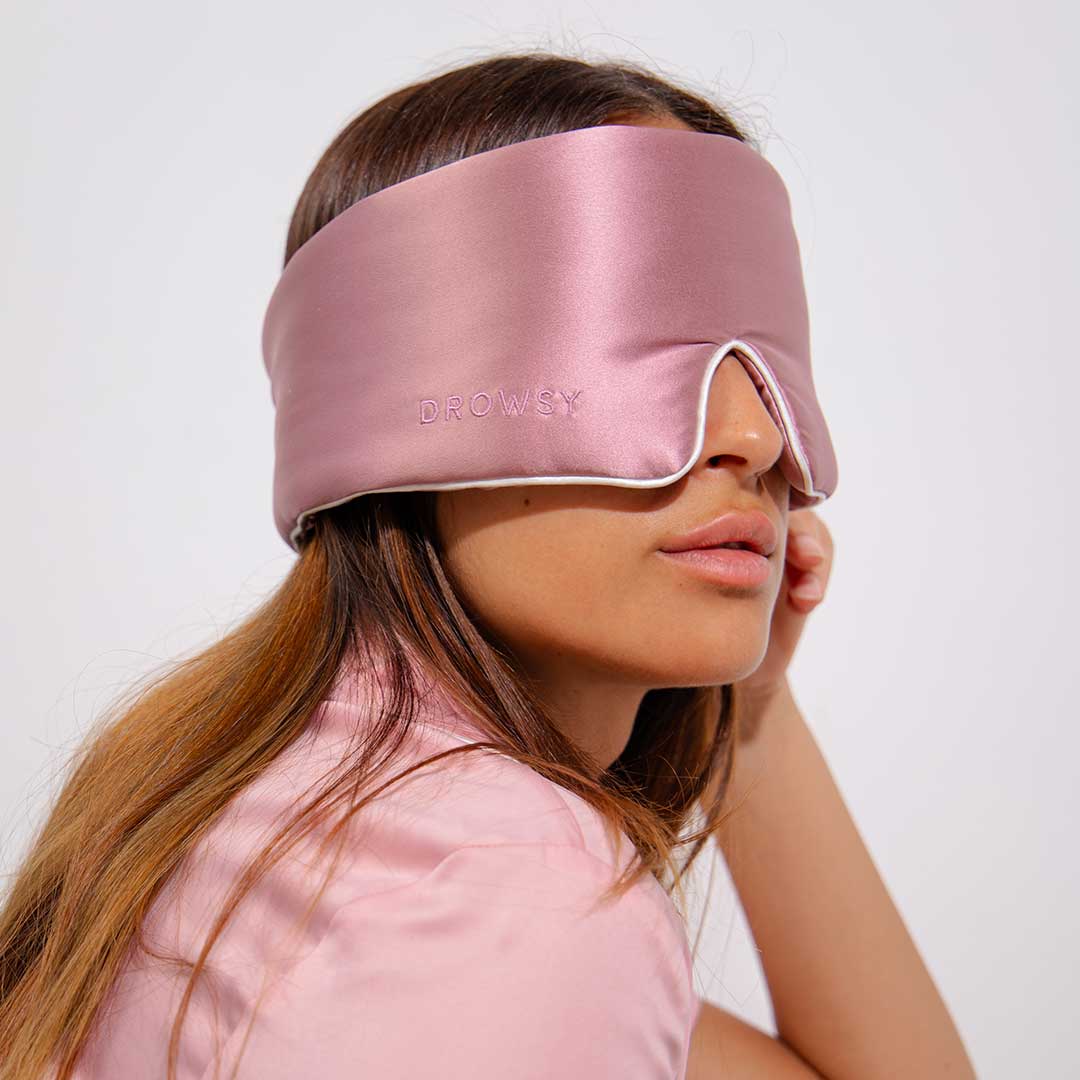 A woman wearing a pink satin sleep mask with the word Drowsy embroidered on it, sitting with her head resting on her hand. She wears a matching pink shirt and has long brown hair.