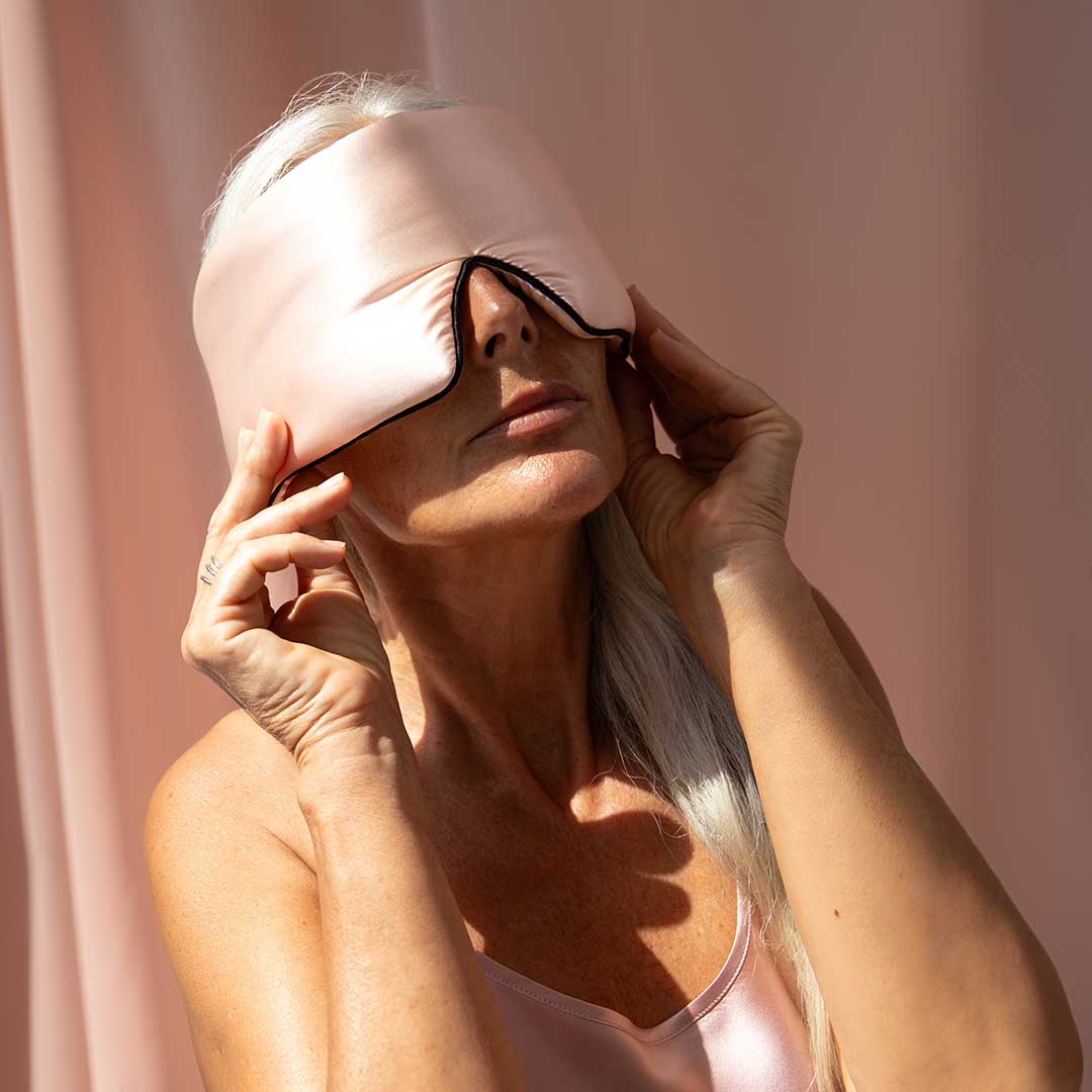 A woman with long silver hair wearing a pink satin sleep mask and matching top adjusts the mask over her eyes. She is standing against a soft pink background in bright, natural light.