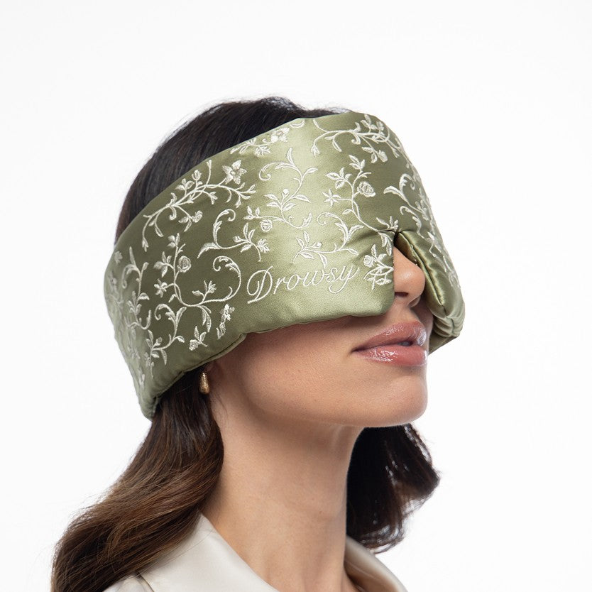 A person wearing a Pride & Prejudice - Sage + English Rose sleep mask in sage green mulberry silk, embroidered with a white floral pattern and the word Drowsy.