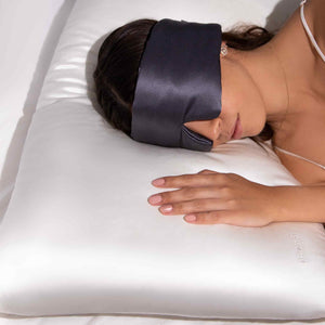 A woman rests on her side with one hand on a white Rajah Cushion Cover by drowsysleepco, wearing a black silk sleep mask and a white strap top. The pillow shows the PILLOW text partially visible.