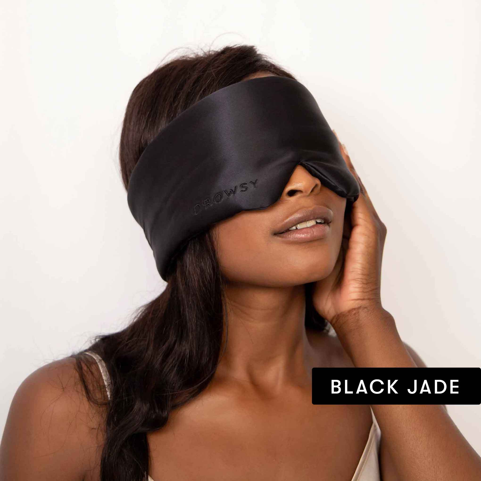 A woman with long dark hair wears the drowsysleepco Lovers Pack luxury silk sleep mask, gently touching her face. The blackout eye mask features DROWSY for comfort and style. The text BLACK JADE appears in the corner.