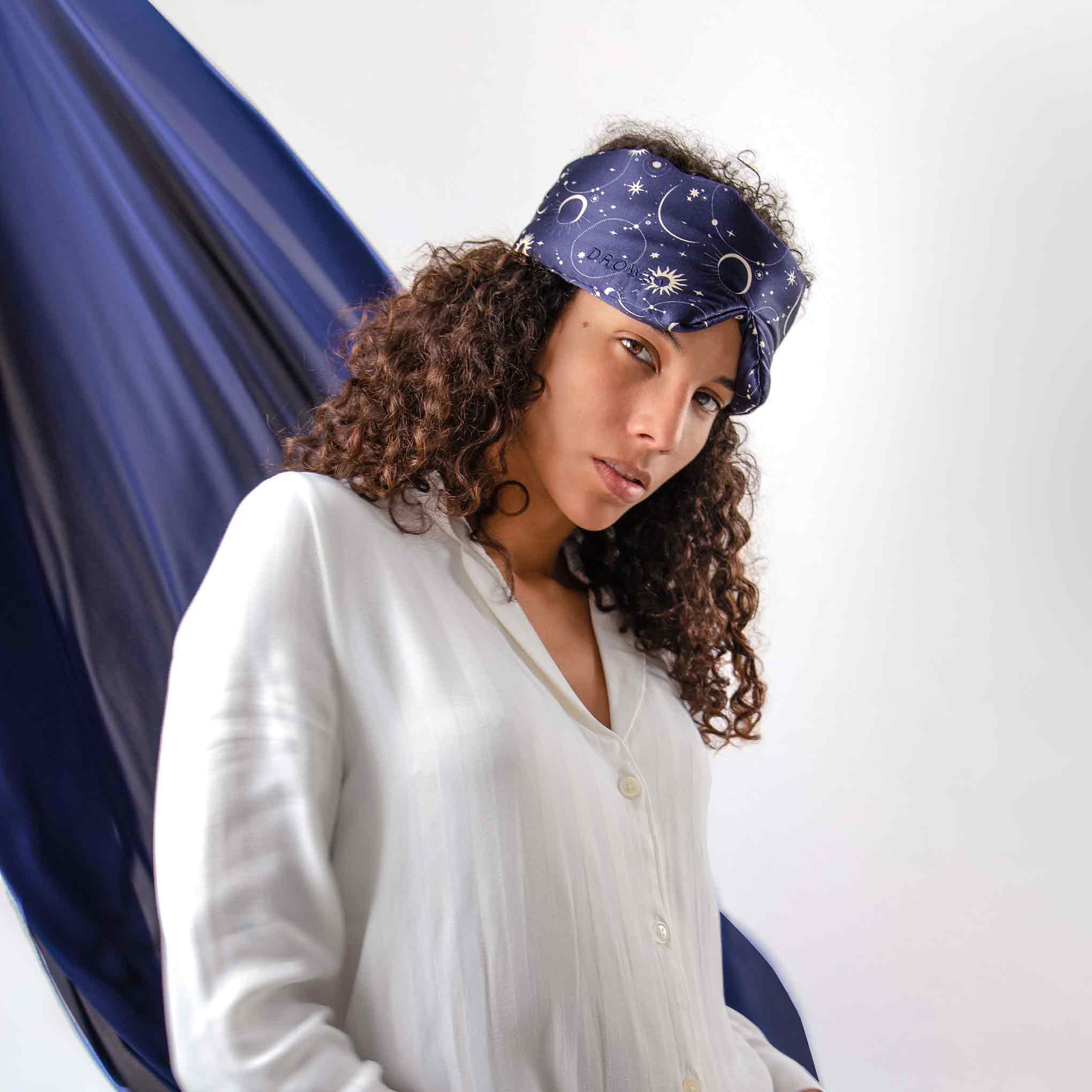 A woman with curly hair wearing a white button-up shirt models the Cosmic Energy silk sleep mask by drowsysleepco, featuring star and moon patterns, standing before a flowing dark blue fabric backdrop.
