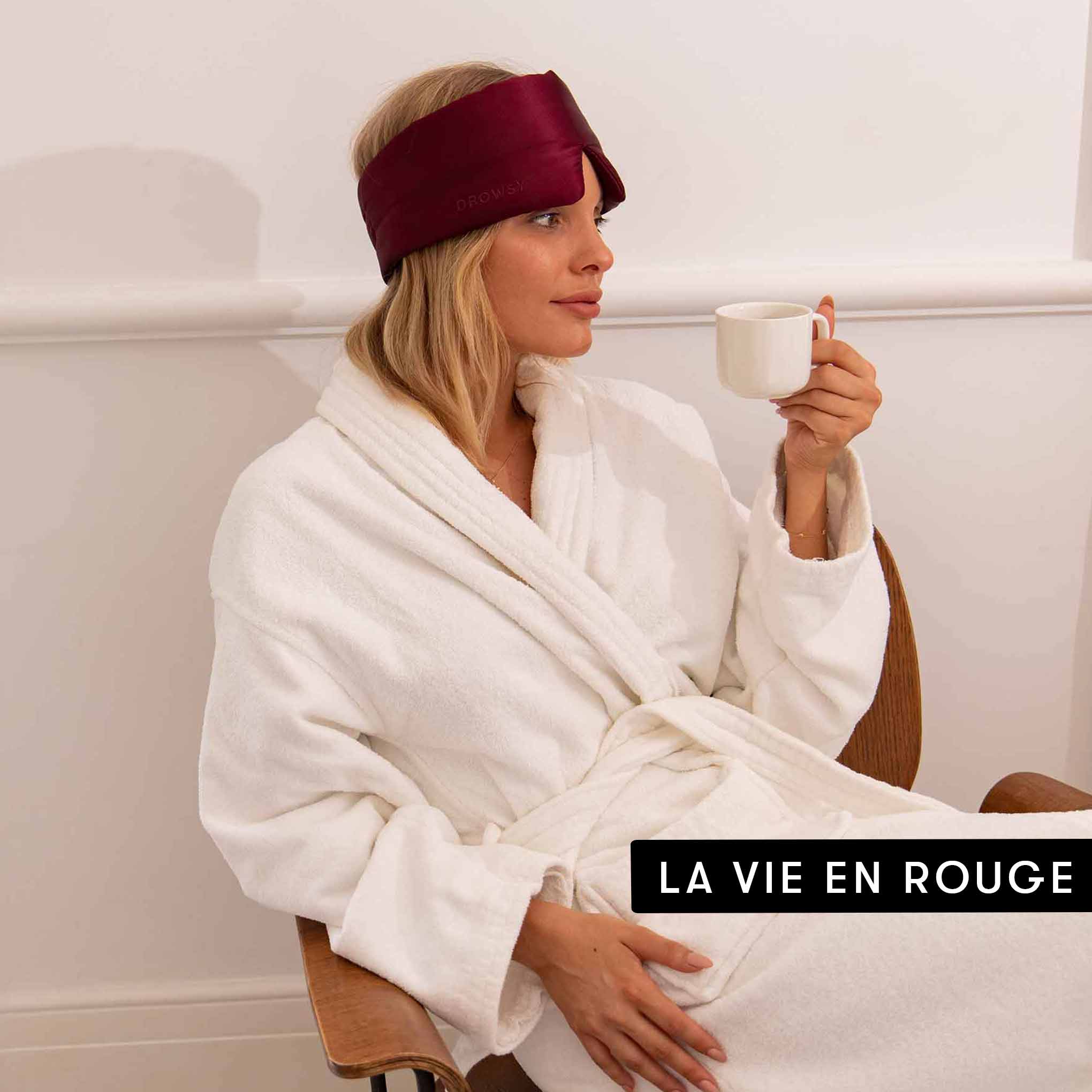 A woman in a white bathrobe and burgundy drowsysleepco Lovers Pack silk sleep mask sits in a chair, holding a white cup. LA VIE EN ROUGE appears in bold text at the lower right.