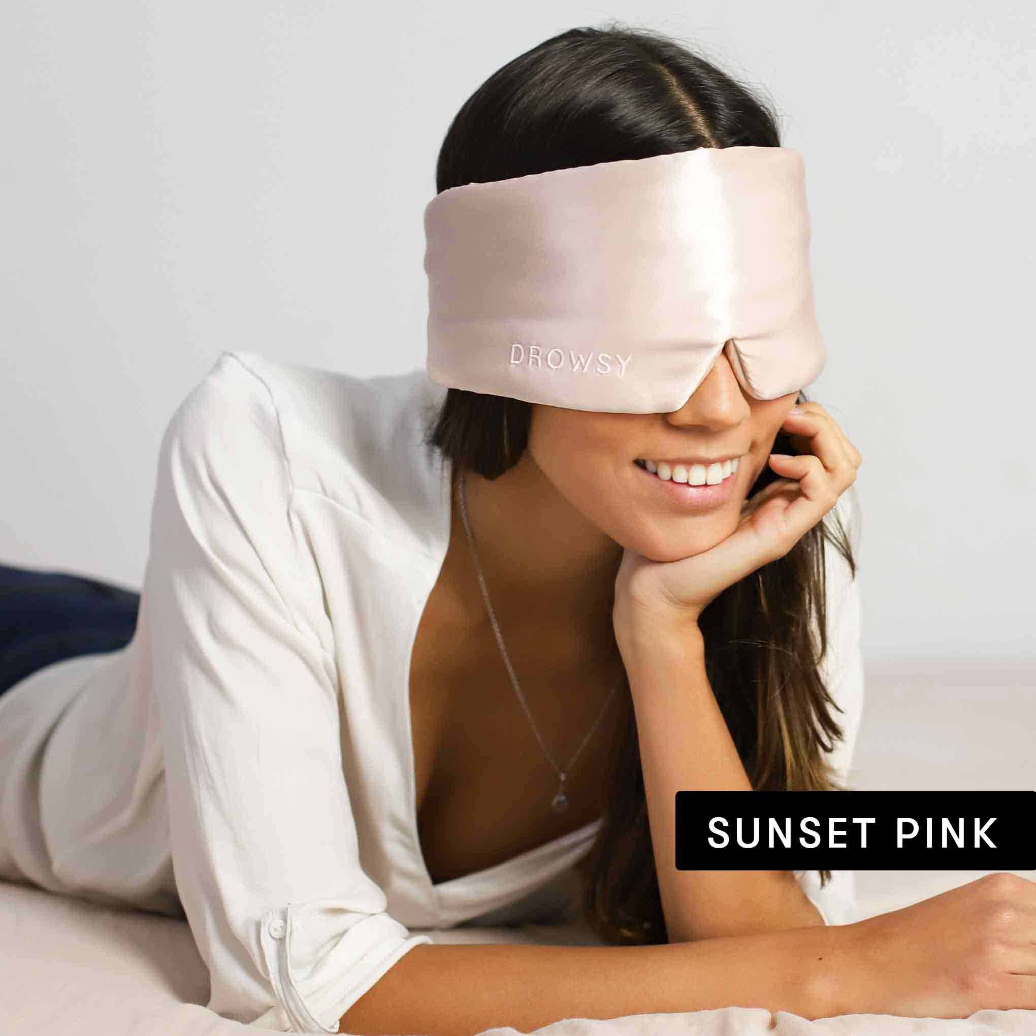 A woman smiles while lying on a bed, wearing a shiny, light pink drowsysleepco sleep mask from the Lovers Pack collection. She’s dressed in white, with SUNSET PINK shown at the bottom right.