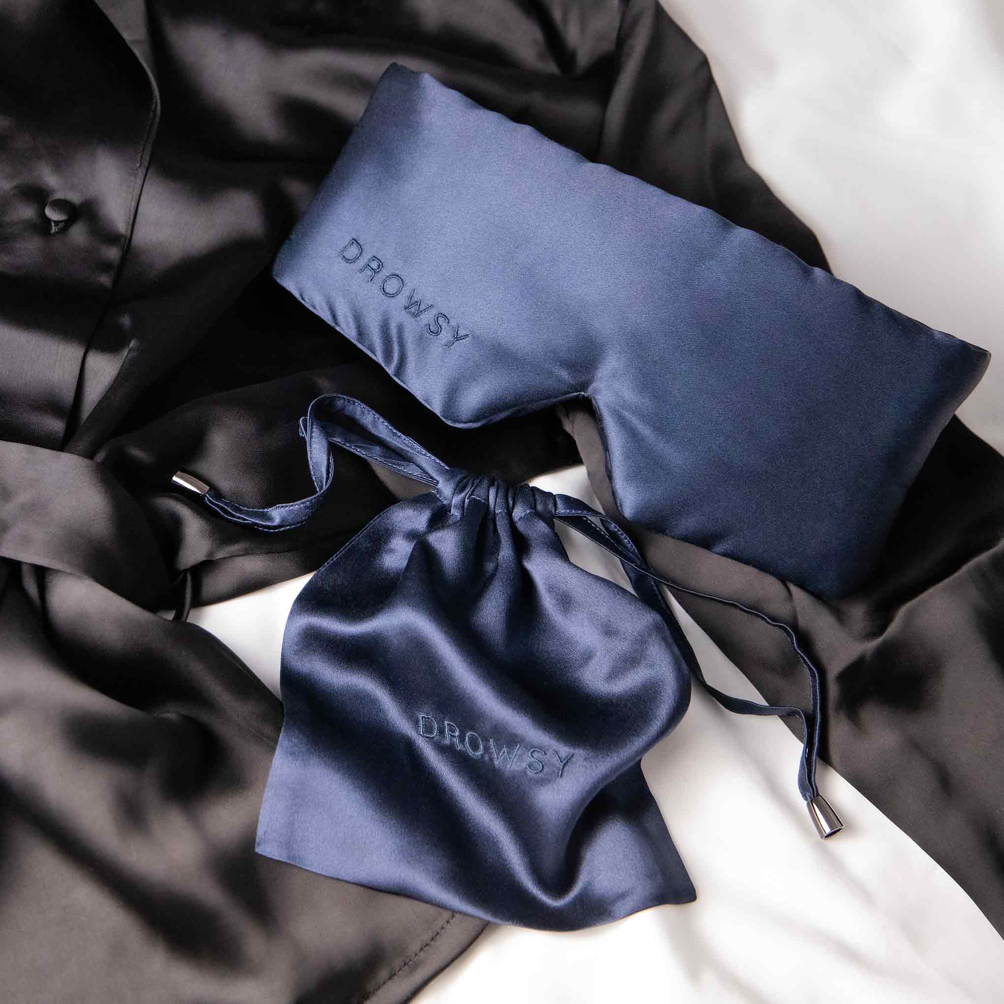 A navy blue silk sleep mask with DROWSY embroidered on it, paired with a matching drawstring pouch, is placed on a black silk robe and white bedsheet.