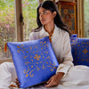 A woman in a white outfit sits indoors by a window, holding the drowsysleepco Magic Carpet Cushion Cover—blue with gold embroidery and tassels. She looks to the side, with another mulberry silk pillow and decor in the background.