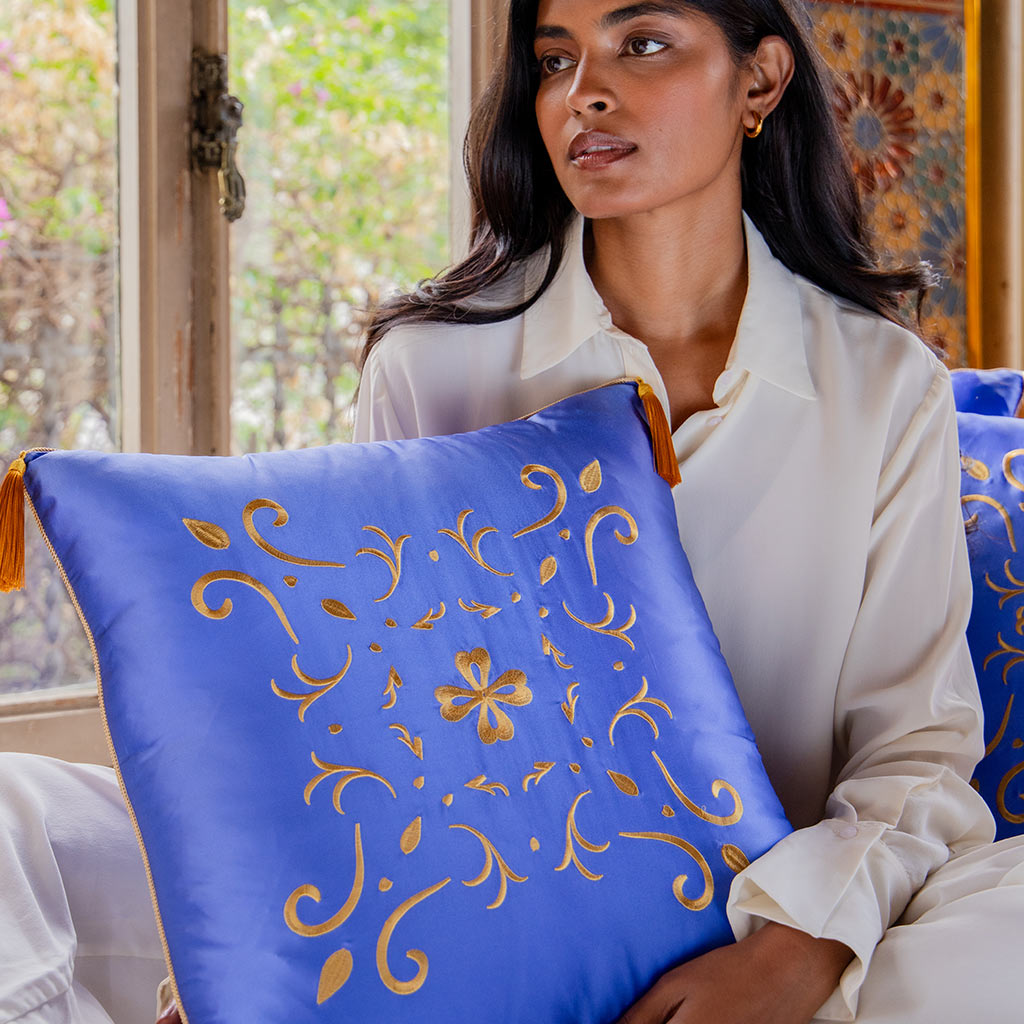 A woman in white sits indoors, holding the drowsysleepco Magic Carpet Cushion Cover—a large blue cushion with gold floral and leaf embroidery. Another matching cushion rests nearby by a window overlooking lush greenery.