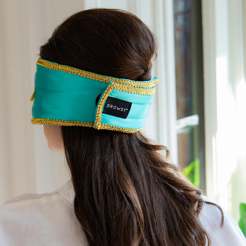 A woman with long brown hair wears a teal, gold-trimmed drowsysleepco 2-Pack Sleep Mask from the Breakfast at Drowsy collection, covering her eyes while standing indoors by a sunlit window.