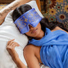 A woman in a blue dress rests on a wooden bed with a colorful mosaic wall, her eyes covered by the drowsysleepco Magic Carpet Sleep Mask, featuring ornate blue and gold mulberry silk.