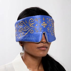 A woman with long dark hair wears the drowsysleepco Magic Carpet Sleep Mask, adorned with blue and gold embroidery. The mask covers her eyes and part of her nose as she pairs it with a white shirt and gold earring.