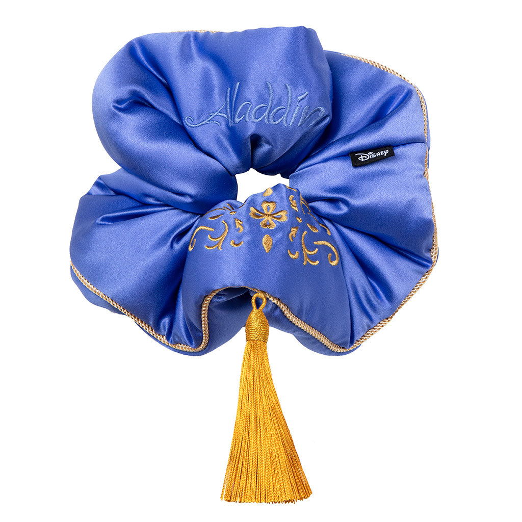 The Magic Carpet Padded Scrunchie by drowsysleepco is a royal blue oversized silk scrunchie with gold embroidery, a gold tassel, "Aladdin" stitched on it, and a Disney tag—designed to boost hair thickness with a magical touch.
