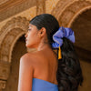 A woman with long, dark hair styled in a ponytail with the drowsysleepco Magic Carpet Padded Scrunchie poses by ornate arches. She wears a blue strapless top and small gold hoops, highlighting her accessory for thicker-looking hair.
