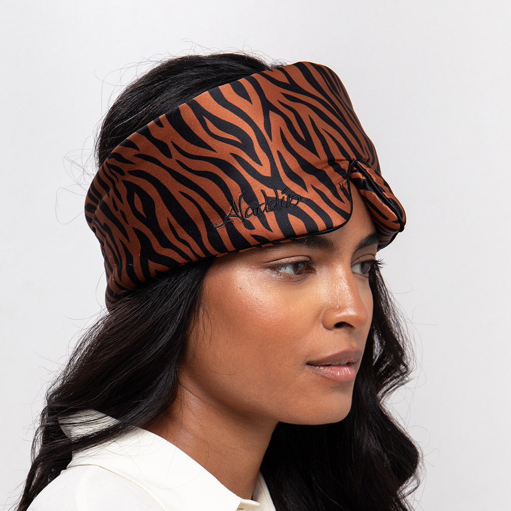 A woman with long dark hair wears the Rajah Sleep Mask by drowsysleepco, featuring a brown and black tiger-striped pattern. She faces slightly sideways, her complexion as fresh and smooth as after a restful night using the mulberry silk mask.