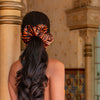 A woman with long wavy dark hair, seen from behind before ornate golden columns, wears the drowsysleepco Rajah Padded Scrunchie in orange and black, highlighting her stylish choice for hair protection.