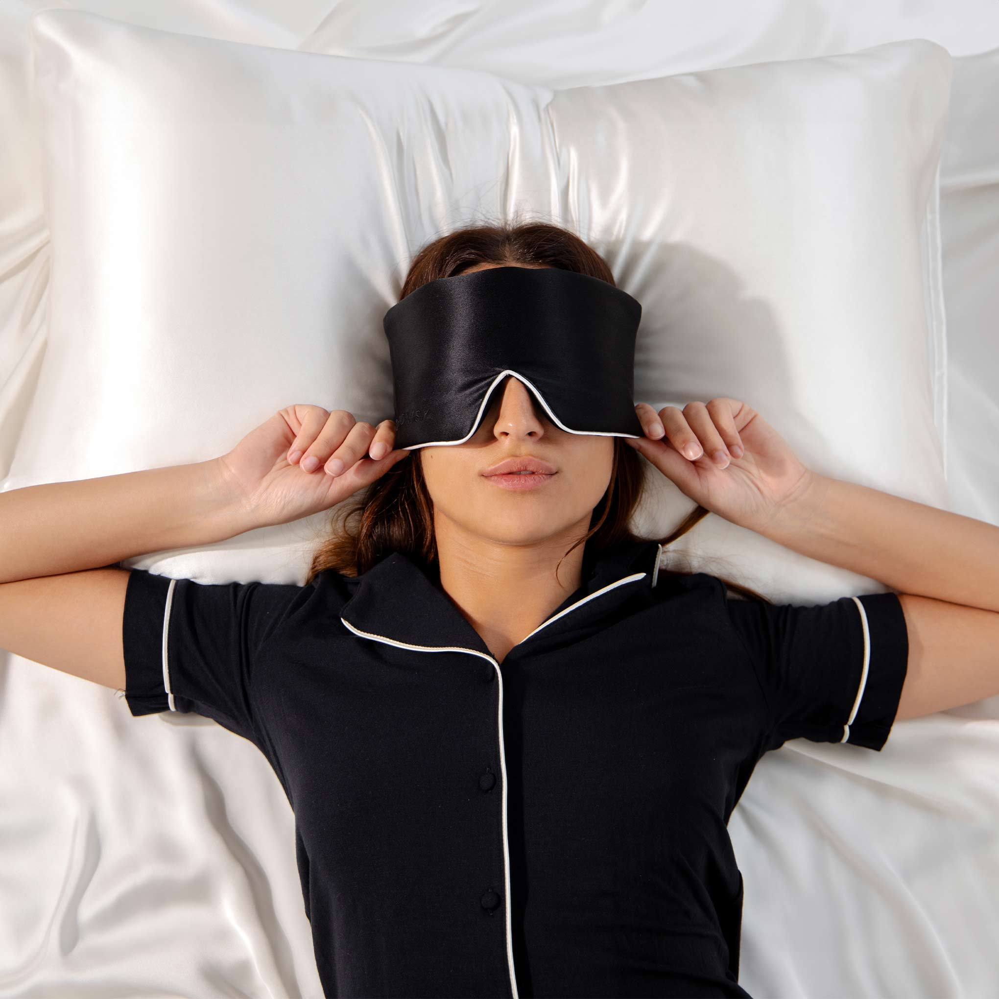 Model lying on white Drowsy silk pillowcase with a black silk sleep mask covering her eyes