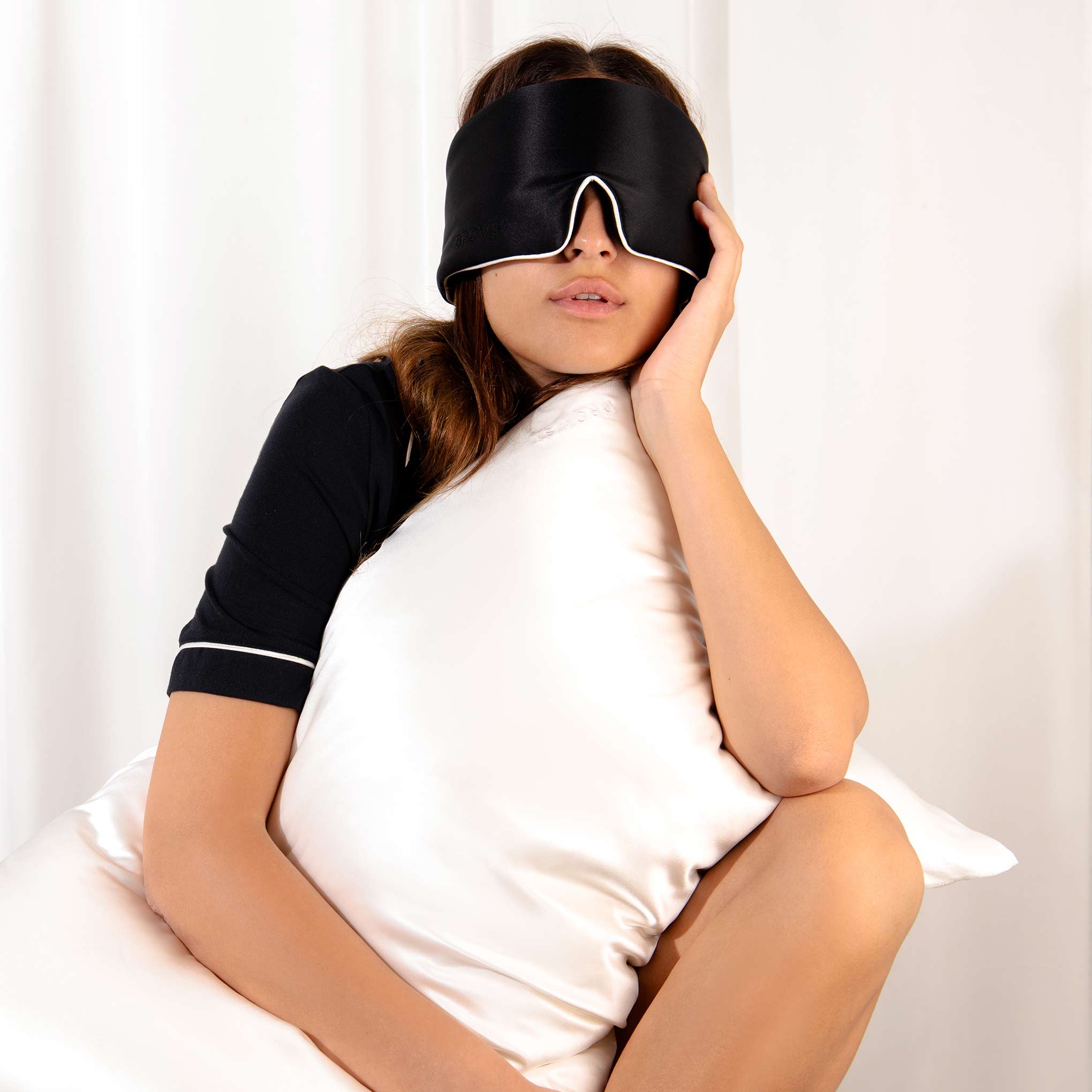 Model hugging white Drowsy silk pillowcase with a black silk sleep mask covering her eyes