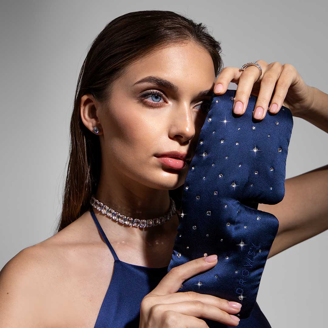 A woman in a satin halter dress holds a dark blue, gem-studded sleep mask to her face. She wears a sparkling choker, earrings, and a ring, with her straight brown hair tucked behind her ear.