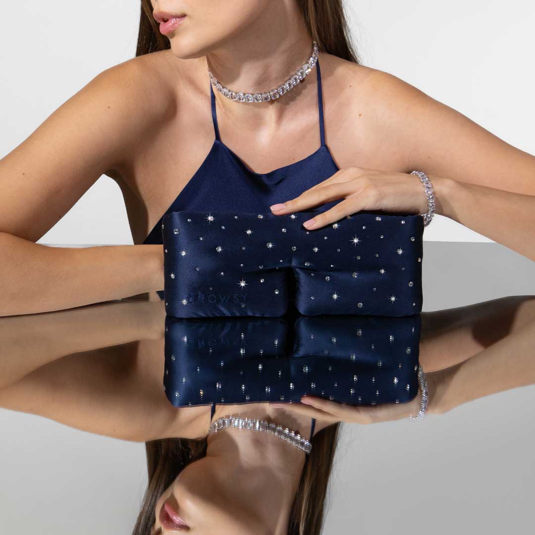 A woman in a blue halter top, wearing a sparkling necklace and bracelet, holds a navy-blue clutch with star-like embellishments, reflected on a shiny surface.