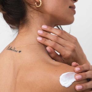 A woman with manicured nails applies drowsysleepco Beauty Sleep Body Cream to her bare shoulder as part of her nighttime ritual, wearing a gold hoop earring, delicate necklace, and showing a cursive tattoo with a heart on her upper back.