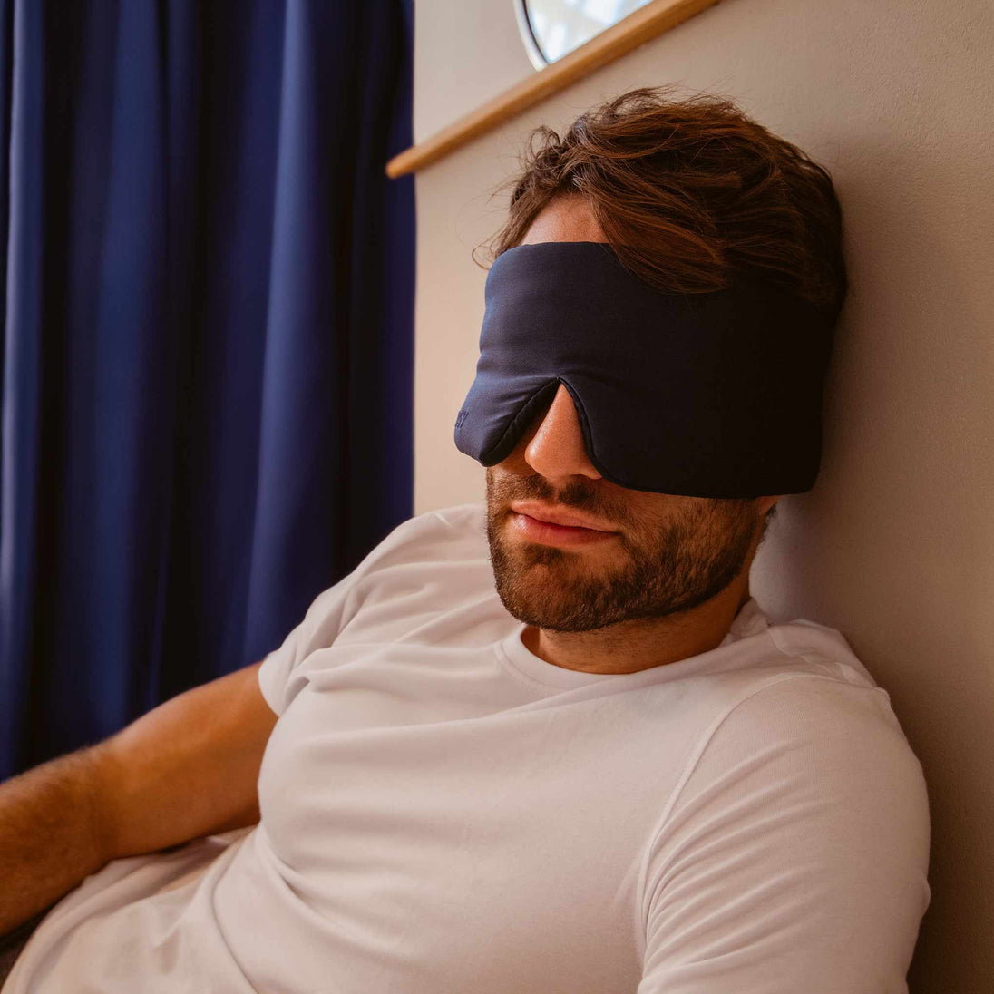 Man leanining against bedroom wall with blue silk curtain in the background and a blue Drowsy silk sleep mask covering his eyes