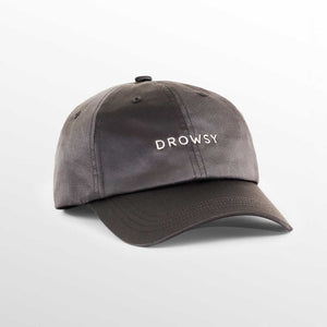 The Drowsy Silk Cap - Moonlight Shadow by Drowsy features DROWSY embroidered in white on gray silk, offering stylish sun protection while keeping your hair flawless, shown against a soft gradient background.