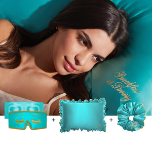 A woman lies on a teal satin pillow from drowsysleepco's Hollywood Experience Bundle, with a matching silk pillowcase, sleep mask, and scrunchie—gold-accented essentials for the ultimate beauty sleep.