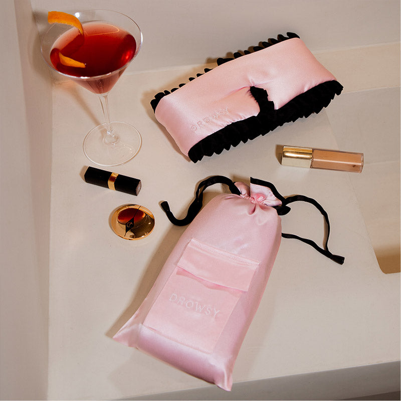 The drowsysleepco Sleep and the City - Pink & Black Sleep Mask + Pouch rests on a cream surface next to chic accessories—a stylish way to improve skin and elevate your night routine.