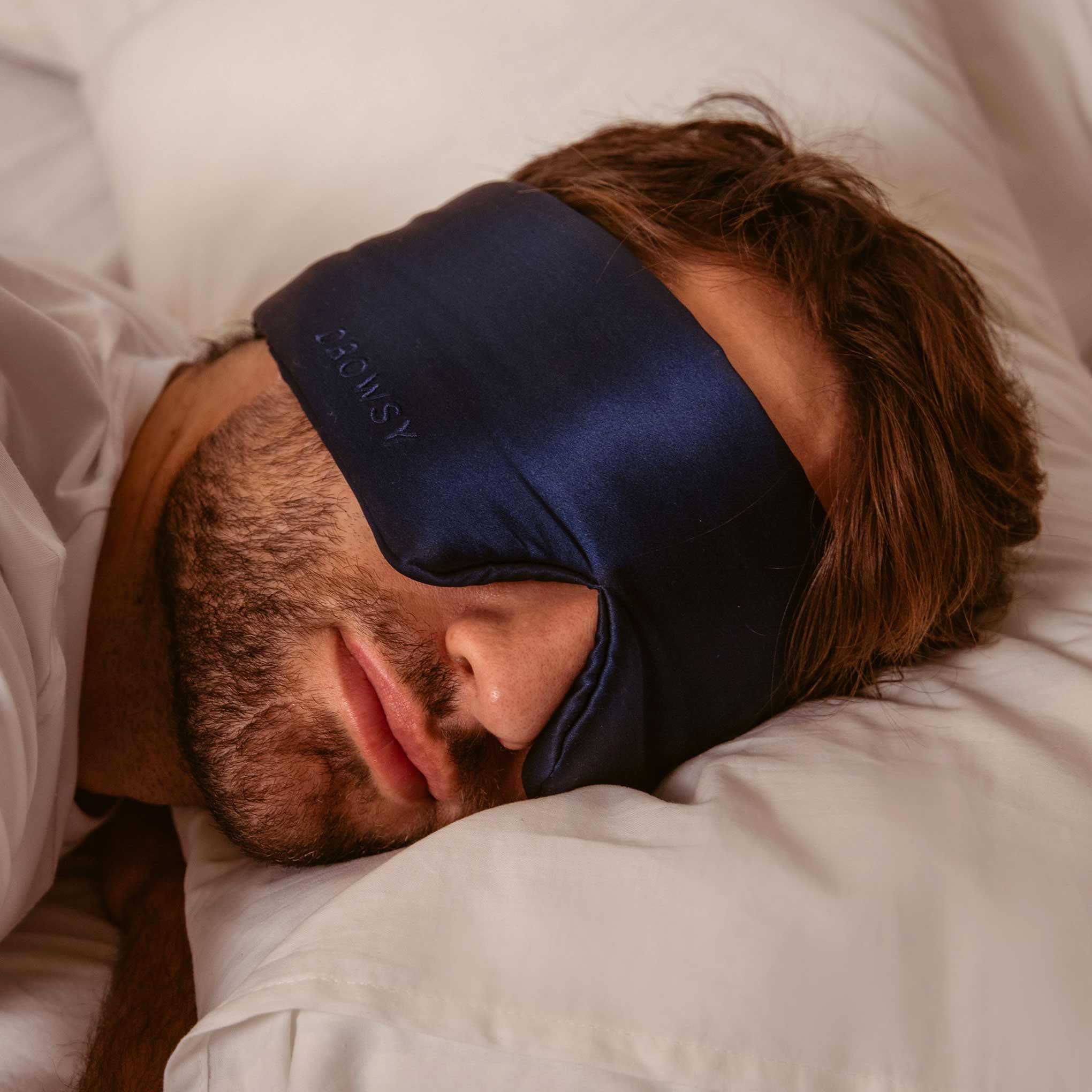 Man sleeping on white bedsheets with a blue Drowsy silk sleep mask covering his eyes