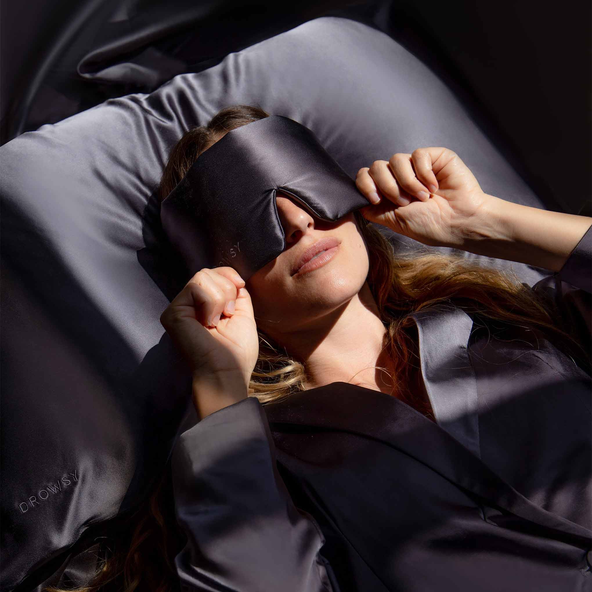 model lying on a charcoal coloured Drowsy silk pillowcase with a charcoal covered Drowsy silk sleep mask covering her eyes