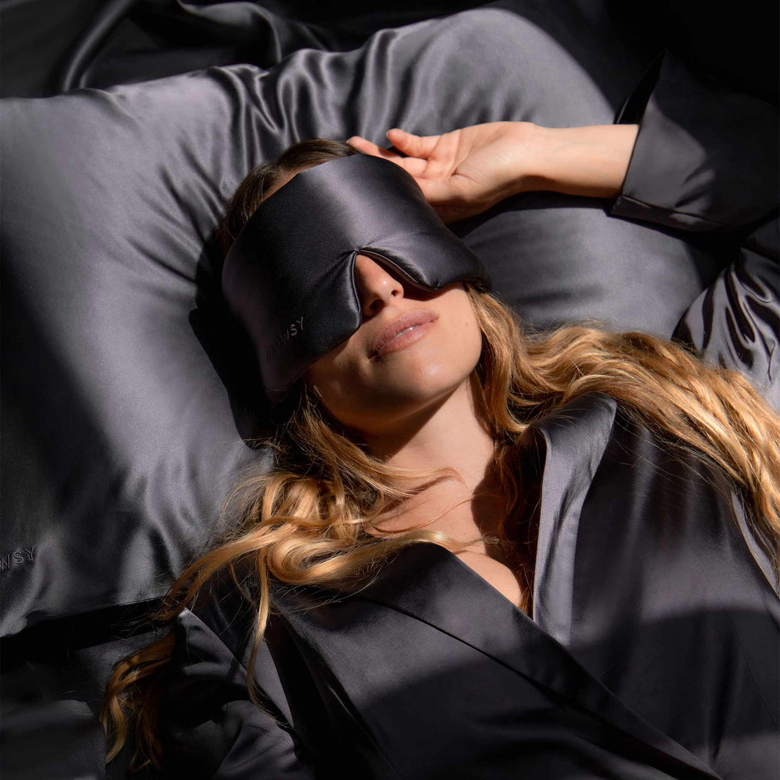 model lying on a charcoal coloured Drowsy silk pillowcase with a charcoal covered Drowsy silk sleep mask covering her eyes