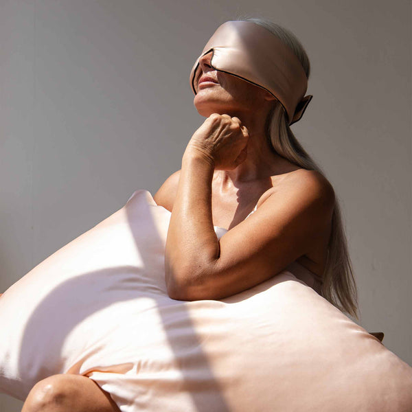 Female model holding a pastel pink coloured Drowsy silk pillowcase with a silk sleep mask covering her eyes