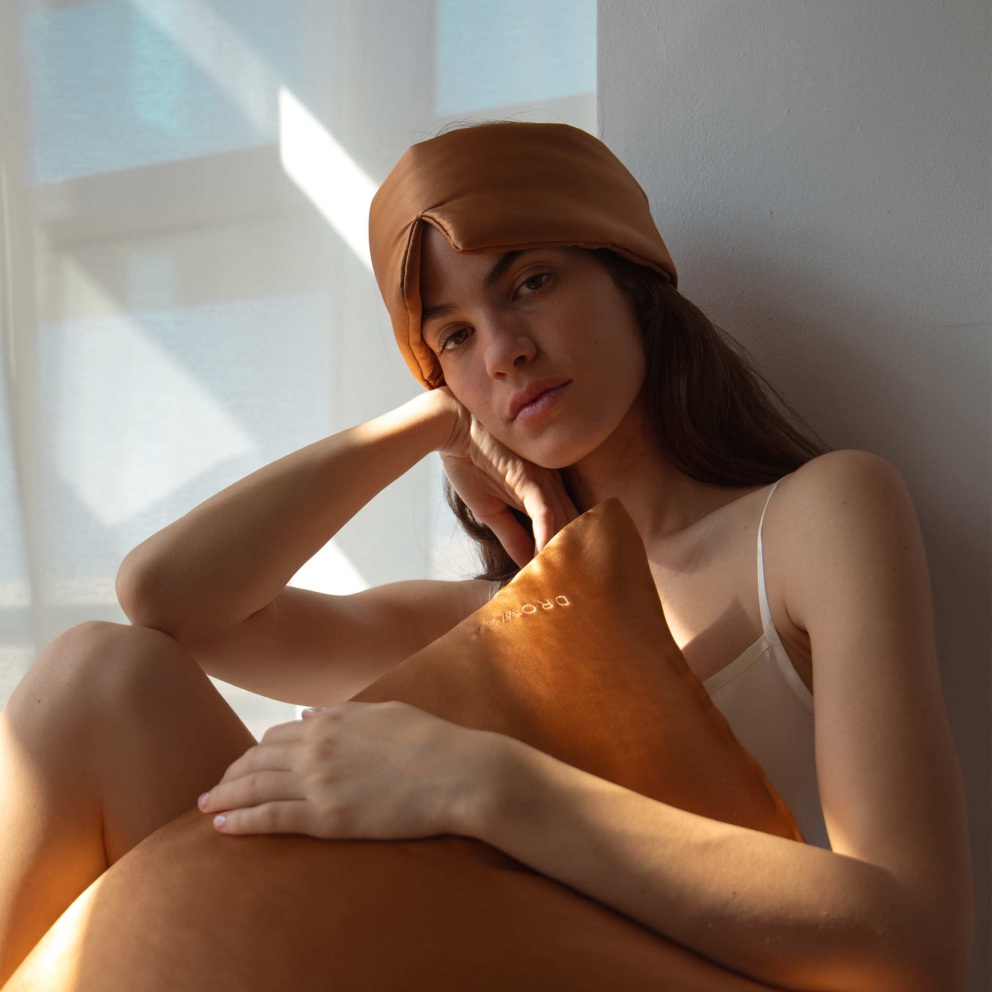 Female model holding terracotta coloured Drowsy silk pillowcase with a Drowsy sleep mask on her forehead