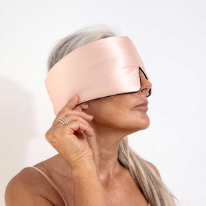 A woman with long gray hair wears the drowsysleepco Sunset Pink - Pipe Dreams Edition eye mask, lightly touching it with her right hand against a plain white background.