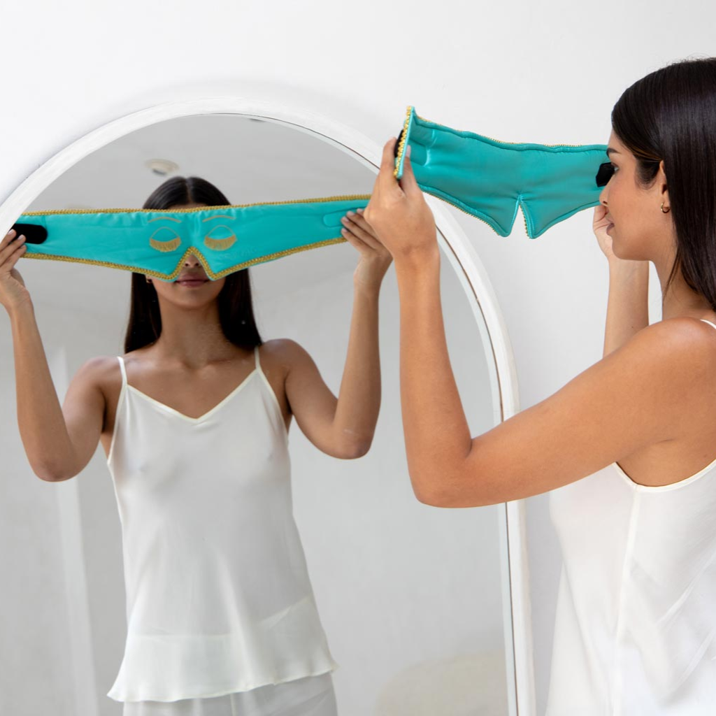 A woman wearing a white camisole stands before a mirror, holding the Perfect Skin Bundle "Breakfast at Drowsy" by drowsysleepco, ready to complete her beauty sleep routine.