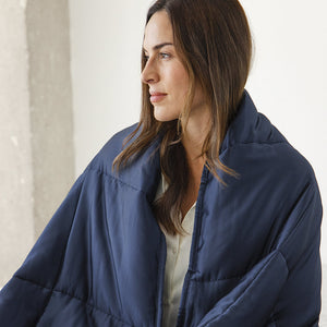 A woman with long brown hair is draped in the drowsysleepco Midnight Blue Silk Weighted Blanket. Indoors and wearing a light shirt, she appears calm and deeply relaxed as she looks to the side.