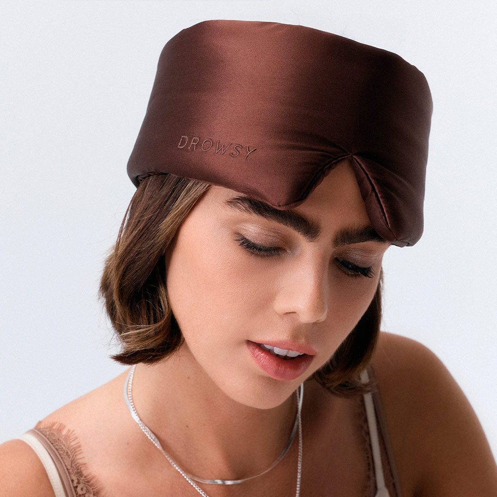 A woman with short brown hair wears a dark brown silk sleep mask labeled "Cocoa Glow" by drowsysleepco, covering her forehead and eyes. She looks neutral, dressed in a tank top and layered necklaces.
