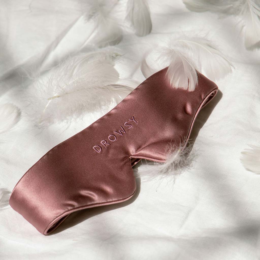 The Damask Rose Featherweight Silk Sleep Mask by drowsysleepco, a silky pink blackout mask embroidered with DROWSY, rests on a white surface amidst soft feathers, as sunlight adds a gentle and peaceful touch.