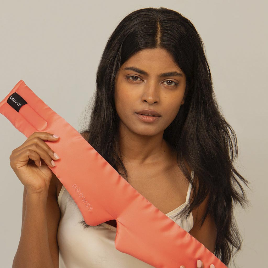 A woman with long dark hair, in a white top, holds the drowsysleepco Summer Coral silk sleep mask against a neutral background, promising the ultimate beauty sleep.