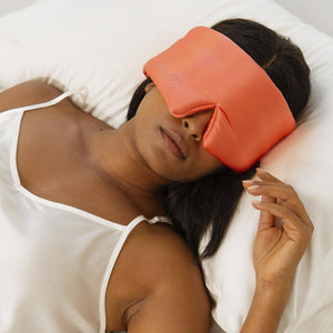 A woman lies on a white pillow wearing drowsysleepcos Summer Coral silk sleep mask, which covers her eyes and nose. Relaxed and on her back in a white satin camisole, she enjoys restful beauty sleep.