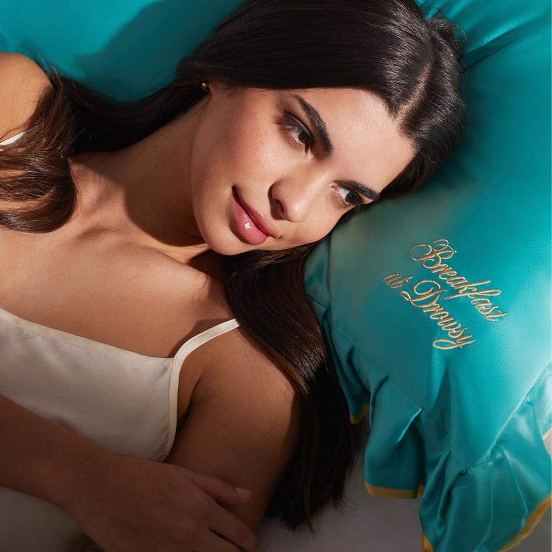 A woman with long dark hair, tied with a silk scrunchie, rests on a turquoise "Breakfast at Dewy" pillow by drowsysleepco, wearing the Hollywood Experience Bundle—an eyelash-protecting accessory—as she smiles softly to the side.