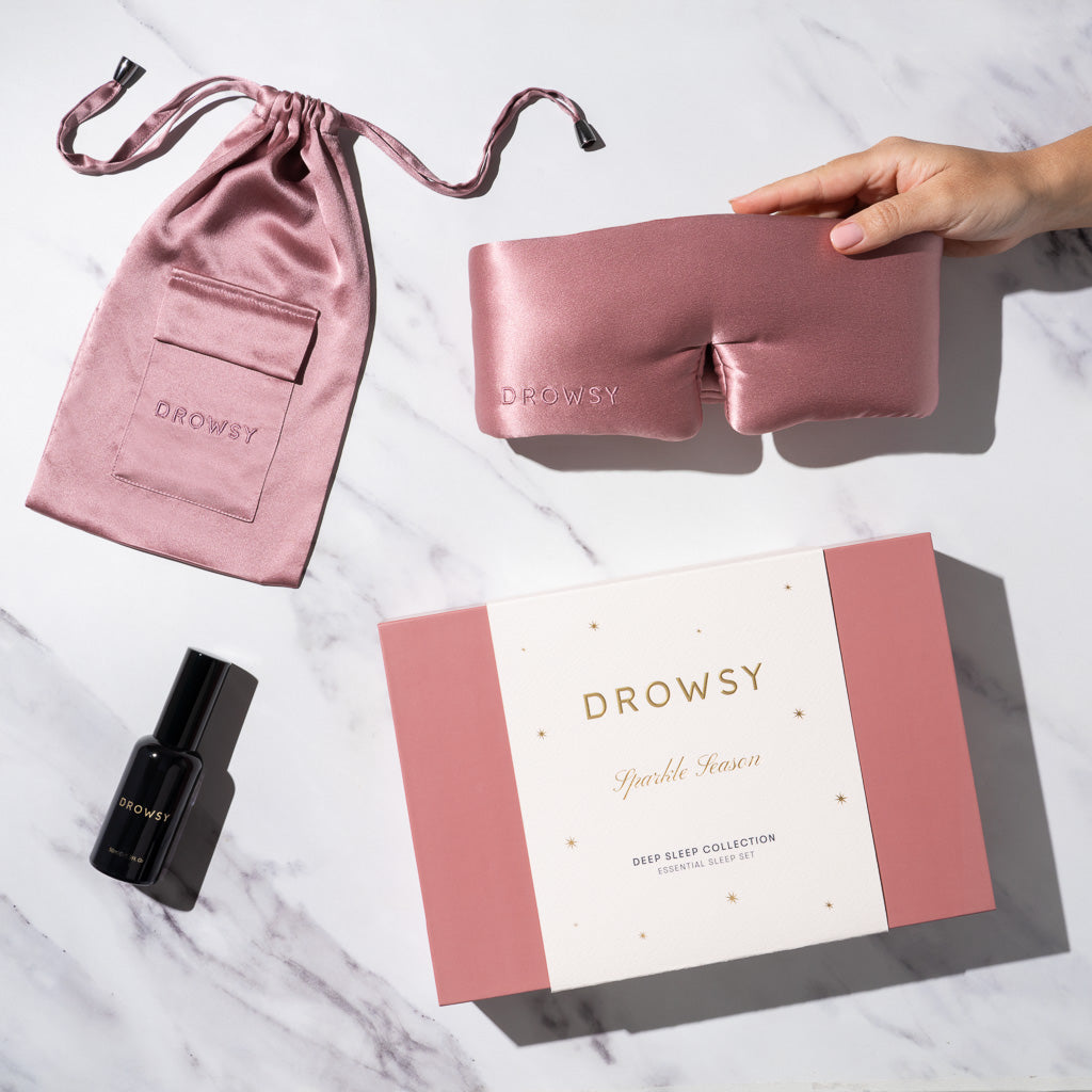 A hand holds a drowsysleepco Damask Rose Deep Sleep Collection silk mask, shown with a pink silk pouch, black deep sleep pillow spray bottle, and pink-and-white drowsysleepco gift box on a white marble surface.