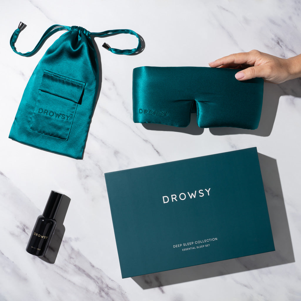 The Green Sapphire Deep Sleep Collection by drowsysleepco sits on a marble surface, with a hand holding the luxury silk sleep mask, alongside a matching silk pouch, deep sleep pillow spray, and a large branded box.
