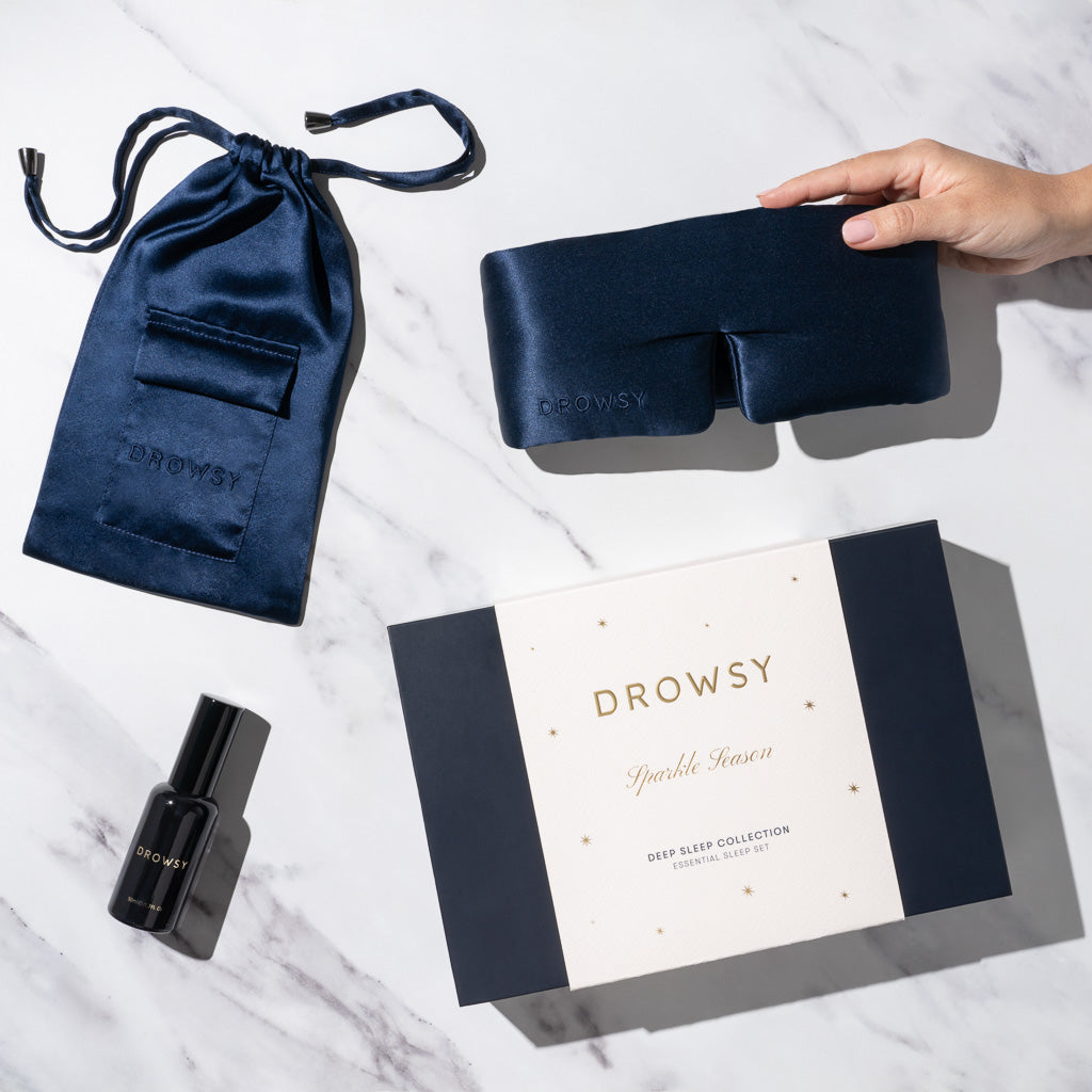 The drowsysleepco Midnight Blue Deep Sleep Collection features a luxury silk sleep mask, matching pouch, DROWSY-labeled pillow spray, and a gift box—elegantly arranged on marble as a hand holds the mask—perfect sleep essentials.