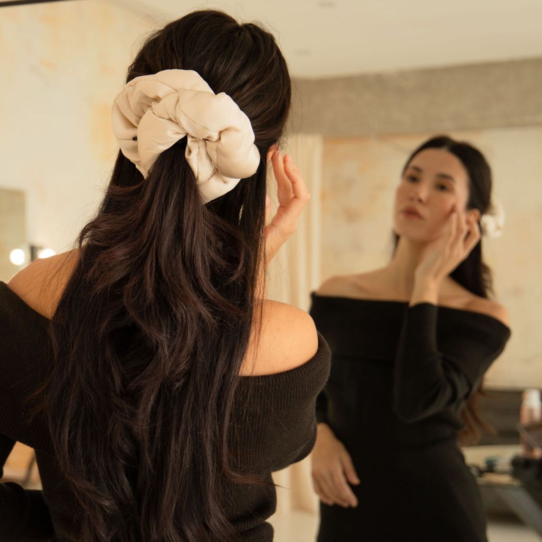 A woman with long dark hair, styled in a large Dusty Gold scrunchie from the Silky Skin & Hair Bundle by drowsysleepco, stands before a mirror in an off-the-shoulder black dress, gently touching her face as she admires her reflection.