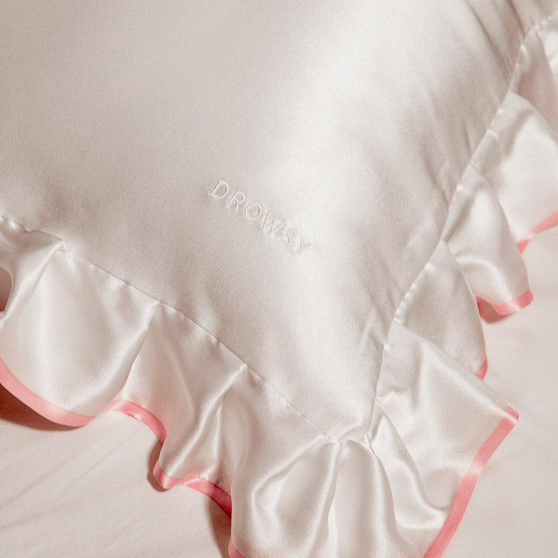 The drowsysleepco Sleep and the City - Pearl & Pink Pillowcase, featuring "DROWSY" embroidered in the center and pink-edged ruffled trim, rests on a bed.
