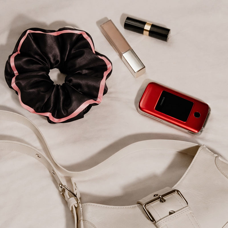 The drowsysleepco Sleep and the City Black Padded Scrunchie, a tube of concealer, lipstick, red flip phone, and a white handbag with a silver buckle are arranged on a light surface.